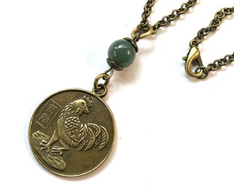 May include: A bronze necklace with a round pendant featuring a rooster design and Chinese characters. The pendant is attached to a chain with a green jade bead.