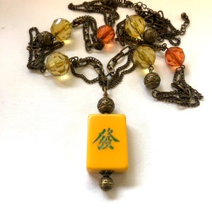Könnte beinhalten: Eine Vintage-Halskette mit einem gelben Anhänger mit grünen chinesischen Schriftzeichen. Die Bronzekette ist mit bernsteinfarbenen und orangefarbenen Perlen verziert. Der rechteckige Anhänger hat Bronzeperlen oben und unten.