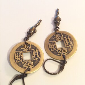 May include: A pair of gold-tone earrings featuring a Chinese coin charm with Chinese characters and a small mouse charm hanging from the top of each coin. The earrings have a hook closure.