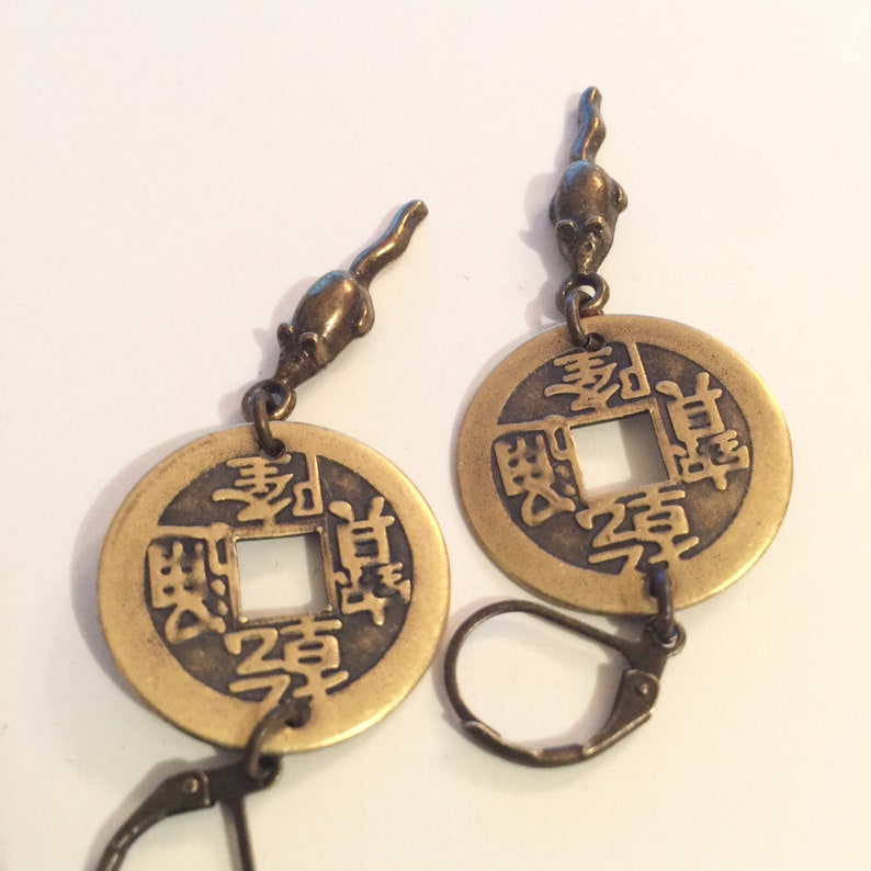 May include: A pair of gold-tone earrings featuring a Chinese coin charm with Chinese characters and a small mouse charm hanging from the top of each coin. The earrings have a hook closure.