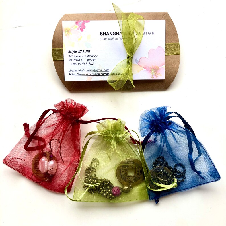 May include: Three small organza bags in red, green, and blue, each containing a different Asian-inspired jewelry piece. The bags are arranged on a white surface. The jewelry includes a pair of earrings, a necklace, and a bracelet.