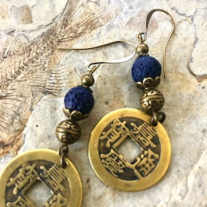 May include: A pair of dangle earrings featuring a brass coin charm with Chinese characters, a dark blue lava rock bead, and a brass bead.
