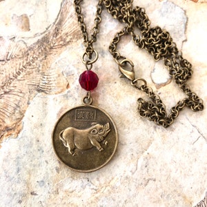 May include: A bronze necklace with a round pendant featuring a pig and Chinese characters. The pendant is attached to a bronze chain with a lobster clasp. A red crystal bead hangs above the pendant.