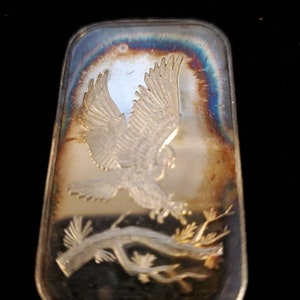 Puede incluir: Barra rectangular de plata con un dise&ntilde;o en relieve de un &aacute;guila en vuelo sobre una rama de &aacute;rbol. La barra tiene una superficie iridiscente con tonos azules, dorados y marrones. Las alas del &aacute;guila est&aacute;n extendidas y los detalles est&aacute;n finamente elaborados.
