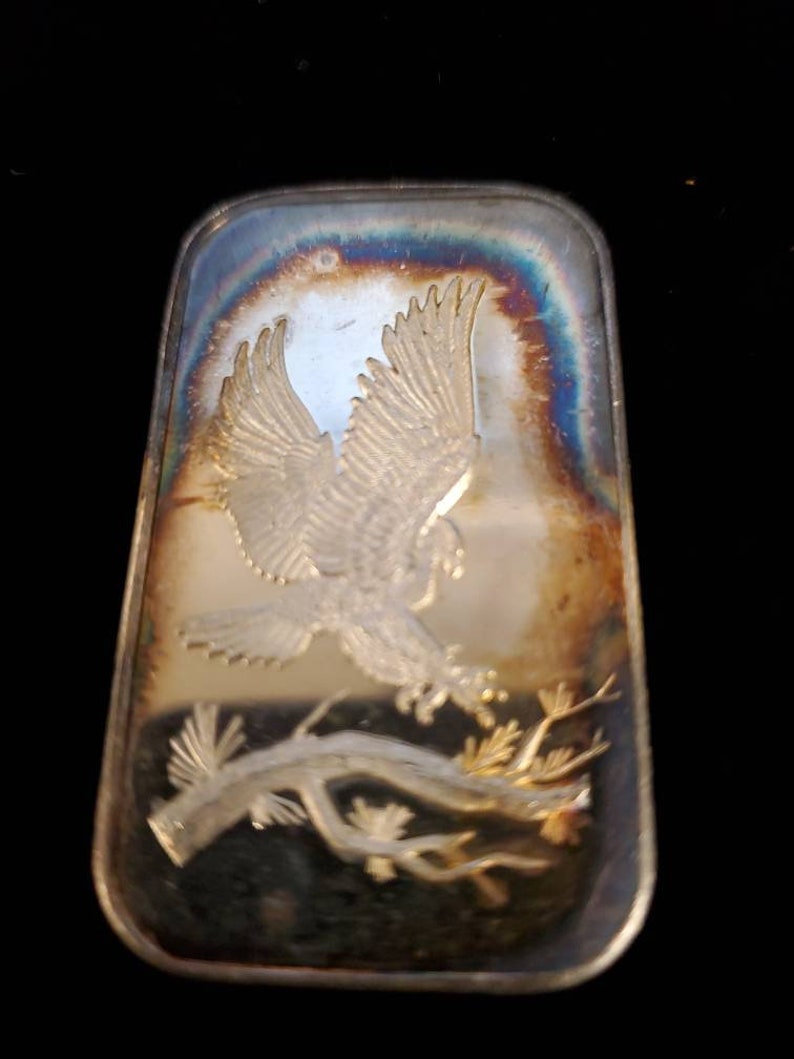 Puede incluir: Barra rectangular de plata con un dise&ntilde;o en relieve de un &aacute;guila en vuelo sobre una rama de &aacute;rbol. La barra tiene una superficie iridiscente con tonos azules, dorados y marrones. Las alas del &aacute;guila est&aacute;n extendidas y los detalles est&aacute;n finamente elaborados.