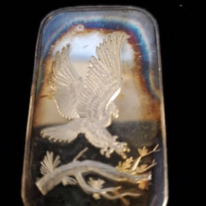 Puede incluir: Barra rectangular de plata con una imagen en relieve de un &aacute;guila en vuelo, agarrando una rama. La barra tiene una superficie reflectante con tonos iridiscentes. El &aacute;guila es el foco central, con patrones detallados de alas y plumas.