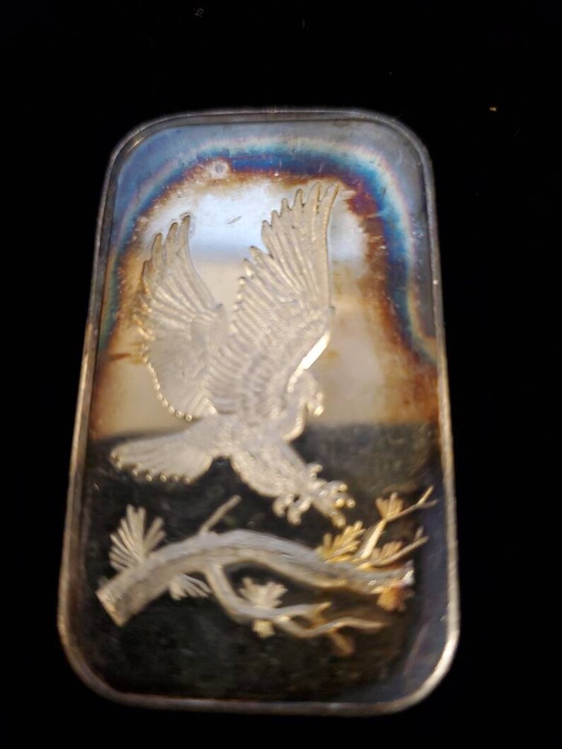 Puede incluir: Barra rectangular de plata con una imagen en relieve de un &aacute;guila en vuelo, agarrando una rama. La barra tiene una superficie reflectante con tonos iridiscentes. El &aacute;guila es el foco central, con patrones detallados de alas y plumas.