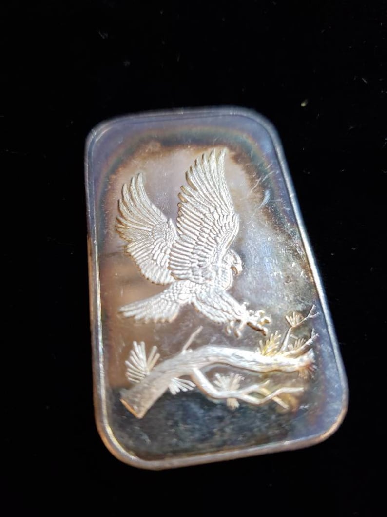 Puede incluir: Barra rectangular de plata con un relieve detallado de un &aacute;guila en vuelo, agarrando una rama. Las alas del &aacute;guila est&aacute;n extendidas y la barra tiene un brillo ligeramente iridiscente. El fondo es negro.