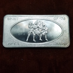 May include: Silver rectangular commemorative coin with the text "SPIRIT OF '76" and "JULY 4 1973". The coin features an oval design with a relief of three figures, one playing a fife and two playing drums, with a flag.