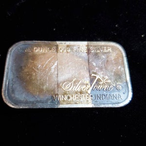 Puede incluir: Barra rectangular de plata con el texto "ONE OUNCE 999 FINE SILVER" y "SilverTowne WINCHESTER, INDIANA". La barra tiene un aspecto deslustrado.