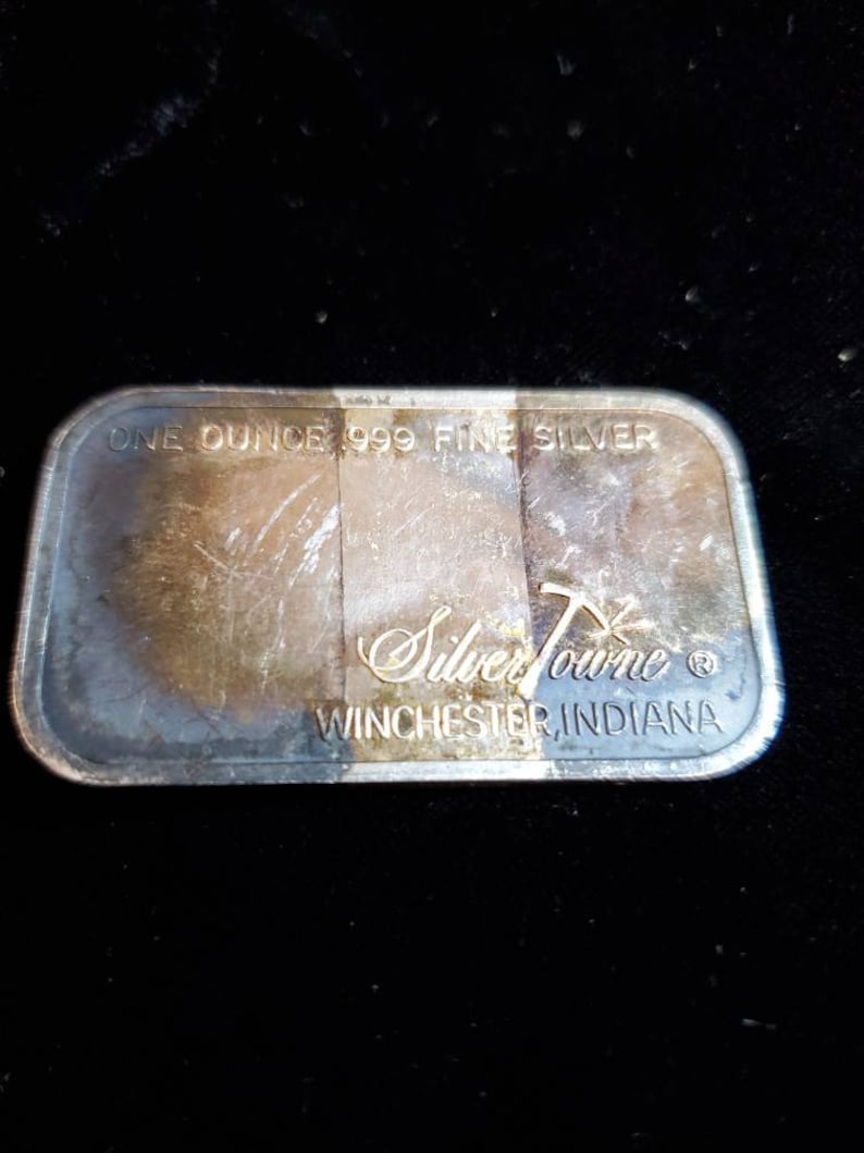 Puede incluir: Barra rectangular de plata con el texto "ONE OUNCE 999 FINE SILVER" y "SilverTowne WINCHESTER, INDIANA". La barra tiene un aspecto deslustrado.