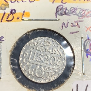 Puede incluir: Una moneda de plata con escritura &aacute;rabe y la fecha 11320c. La moneda est&aacute; en un soporte de moneda de pl&aacute;stico con fondo blanco.