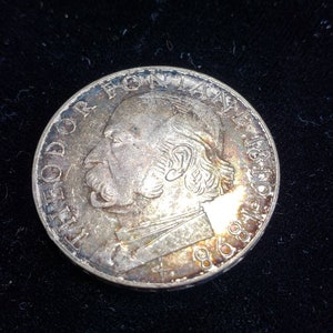 May include: A circular silver-toned commemorative coin featuring a profile portrait of Theodor Fontane facing left. The text THEODOR FONTANE 1819-1898 is embossed along the upper and right edge of the coin, with a small cross symbol at the bottom.