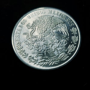 Op de afbeelding: Een zilveren munt met de woorden "ESTADOS UNIDOS MEXICANOS" langs de rand. De munt toont een adelaar met een slang in zijn bek, een symbool van Mexico, omringd door een lauwerkrans.