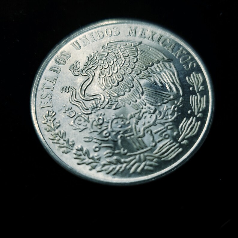Op de afbeelding: Een zilveren munt met de woorden "ESTADOS UNIDOS MEXICANOS" langs de rand. De munt toont een adelaar met een slang in zijn bek, een symbool van Mexico, omringd door een lauwerkrans.