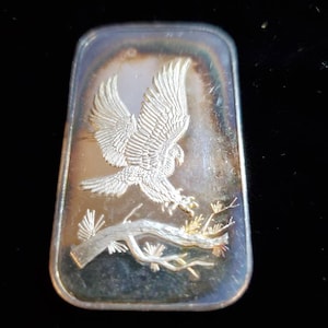 Puede incluir: Barra rectangular de plata con un relieve detallado de un &aacute;guila en vuelo, agarrando una rama. Las alas del &aacute;guila est&aacute;n extendidas y la barra tiene un brillo ligeramente iridiscente. El dise&ntilde;o est&aacute; sobre un fondo oscuro.