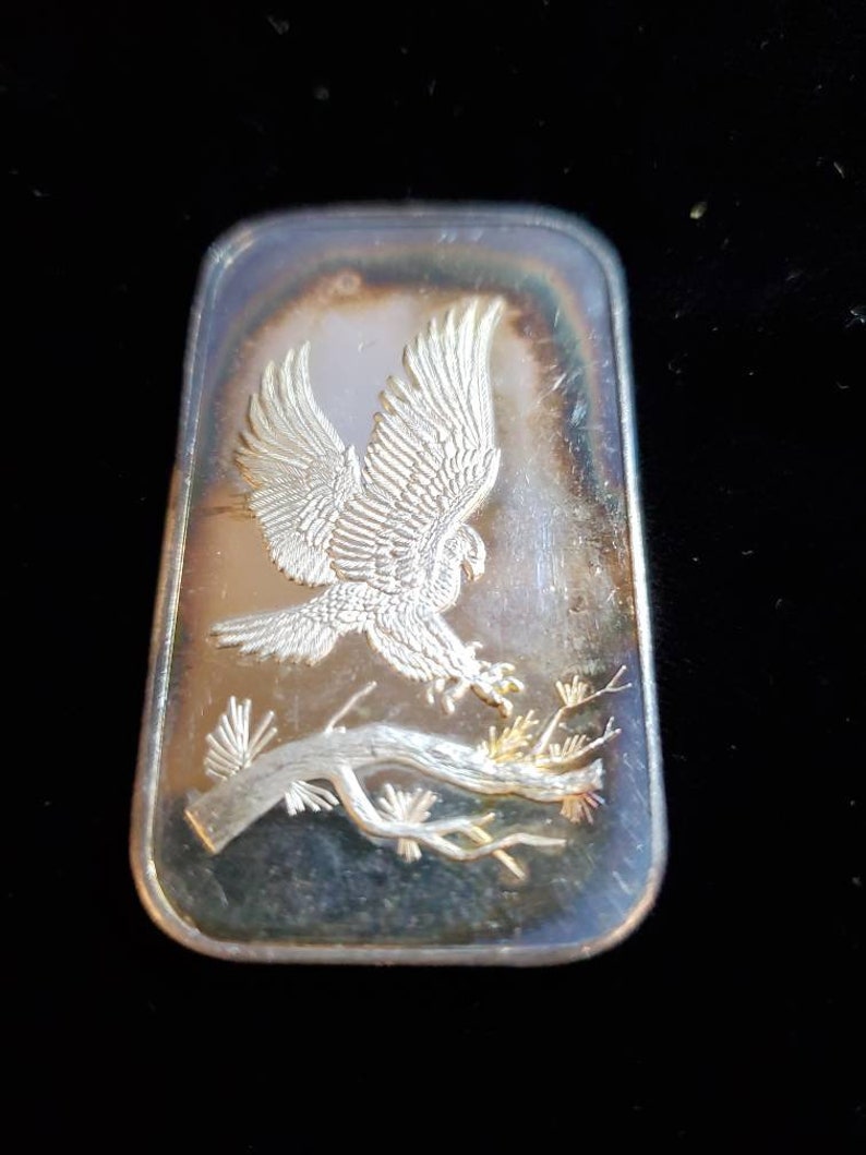 Puede incluir: Barra rectangular de plata con un relieve detallado de un &aacute;guila en vuelo, agarrando una rama. Las alas del &aacute;guila est&aacute;n extendidas y la barra tiene un brillo ligeramente iridiscente. El dise&ntilde;o est&aacute; sobre un fondo oscuro.