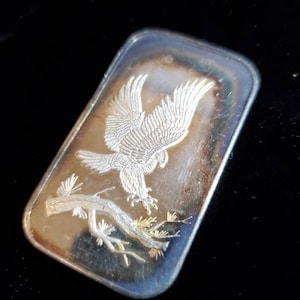 Puede incluir: Barra rectangular de plata con una imagen grabada de un &aacute;guila con las alas extendidas posada en una rama de &aacute;rbol. La barra tiene un acabado pulido con matices iridiscentes en los bordes.