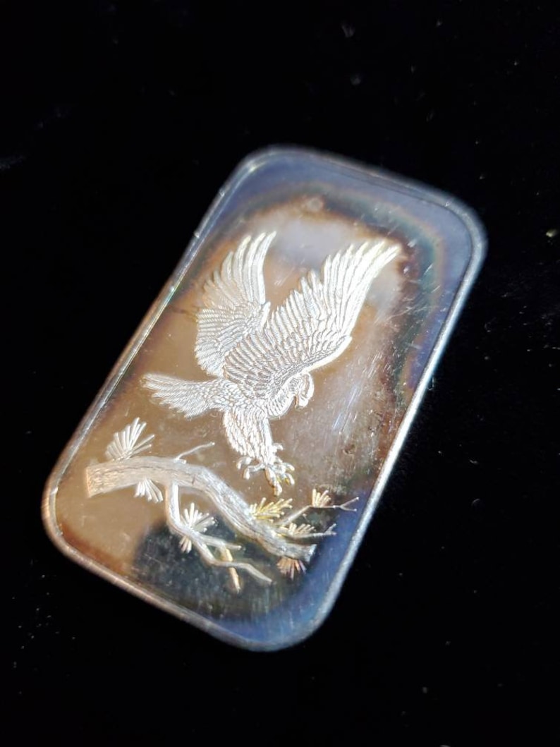 Puede incluir: Barra rectangular de plata con una imagen grabada de un &aacute;guila con las alas extendidas posada en una rama de &aacute;rbol. La barra tiene un acabado pulido con matices iridiscentes en los bordes.