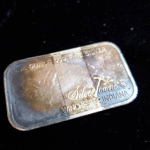 Puede incluir: Barra rectangular de plata con el texto "ONE OUNCE .999 FINE SILVER" y "SilverTowne Winchester, Indiana". La barra tiene un aspecto ligeramente deslustrado.