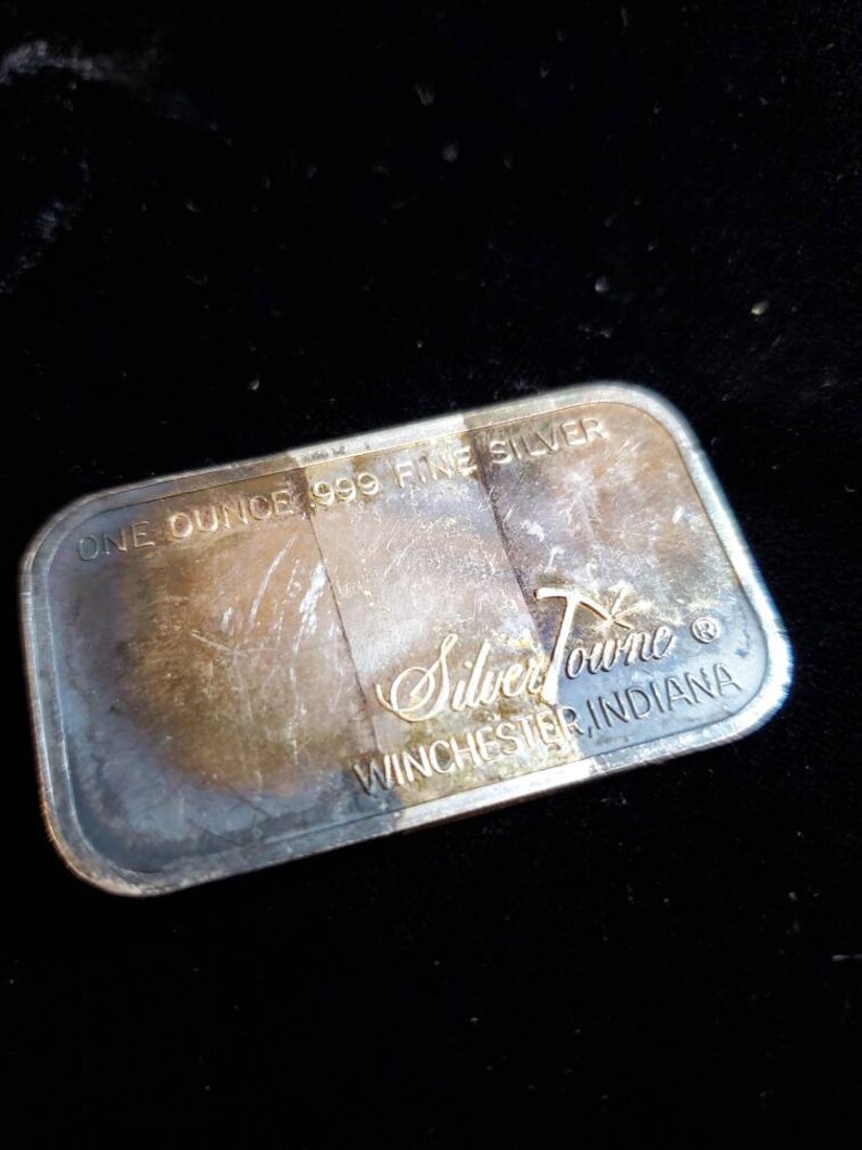 Puede incluir: Barra rectangular de plata con el texto "ONE OUNCE .999 FINE SILVER" y "SilverTowne Winchester, Indiana". La barra tiene un aspecto ligeramente deslustrado.