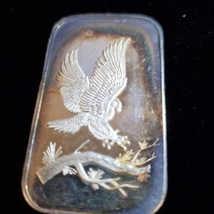 Puede incluir: Barra rectangular de plata con un relieve detallado de un &aacute;guila en vuelo, posada en una rama con follaje. Las alas del &aacute;guila est&aacute;n extendidas y la barra tiene una superficie reflectante.