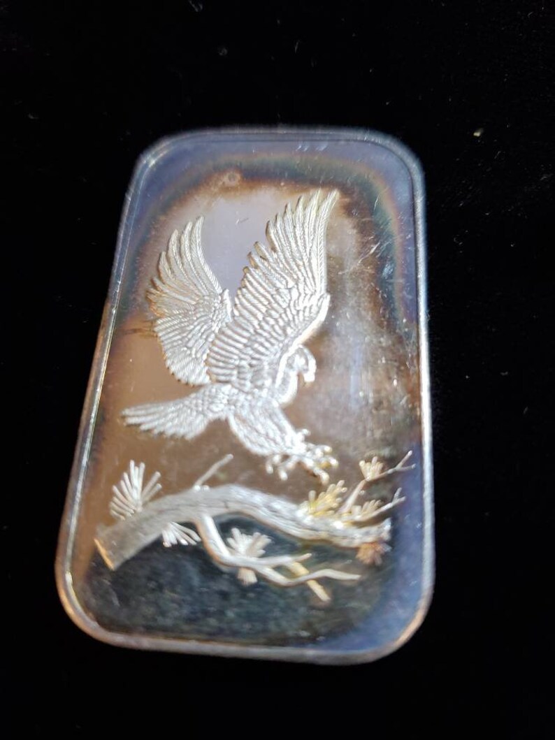Puede incluir: Barra rectangular de plata con un relieve detallado de un &aacute;guila en vuelo, posada en una rama con follaje. Las alas del &aacute;guila est&aacute;n extendidas y la barra tiene una superficie reflectante.