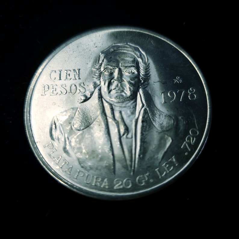 Op de afbeelding: Een zilveren munt met een portret van een man, met de tekst "CIEN PESOS" en het jaartal "1978". De munt bevat ook de tekst "PLATA PURA 20 Gr. LEY .720". De munt staat tegen een zwarte achtergrond.