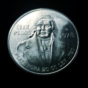 Op de afbeelding: Een zilveren munt met een portret van een man, met de tekst "CIEN PESOS" en het jaartal "1978". De munt bevat ook de tekst "PLATA PURA 20 G. LEY .720". De achtergrond is zwart.