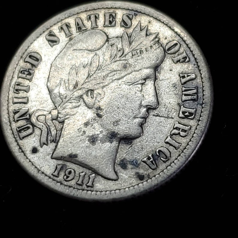 Peut inclure: Une pi&egrave;ce de monnaie am&eacute;ricaine en argent de dix cents avec l'ann&eacute;e 1911 inscrite en bas. La pi&egrave;ce repr&eacute;sente un portrait de la Libert&eacute; avec une couronne de laurier sur la t&ecirc;te et les mots "UNITED STATES OF AMERICA" autour du bord.
