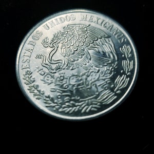 Op de afbeelding: Een zilveren munt met de tekst "ESTADOS UNIDOS MEXICANOS" langs de rand. De munt toont een adelaar met een slang in zijn bek, een Mexicaans symbool, omringd door een lauwerkrans.