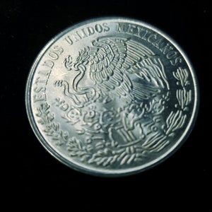 Op de afbeelding: Een close-up van een zilveren munt met de woorden "ESTADOS UNIDOS MEXICANOS" rond de rand. De munt toont een gedetailleerd embleem van een adelaar met een slang in zijn bek, omringd door bladeren.