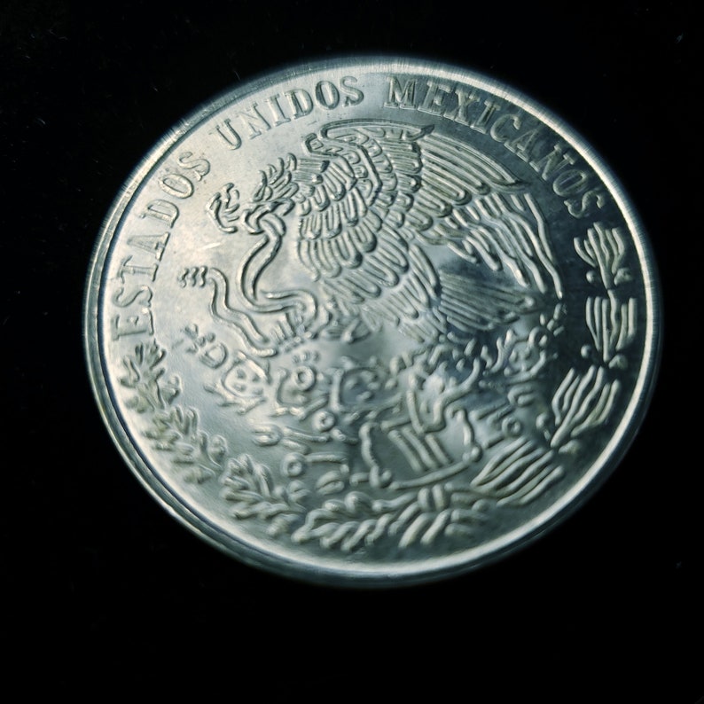 Op de afbeelding: Een close-up van een zilveren munt met de woorden "ESTADOS UNIDOS MEXICANOS" rond de rand. De munt toont een gedetailleerd embleem van een adelaar met een slang in zijn bek, omringd door bladeren.