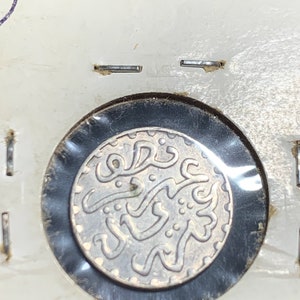 Puede incluir: Una moneda de plata con escritura &aacute;rabe y un dise&ntilde;o decorativo. La moneda est&aacute; encerrada en un soporte de pl&aacute;stico transparente.