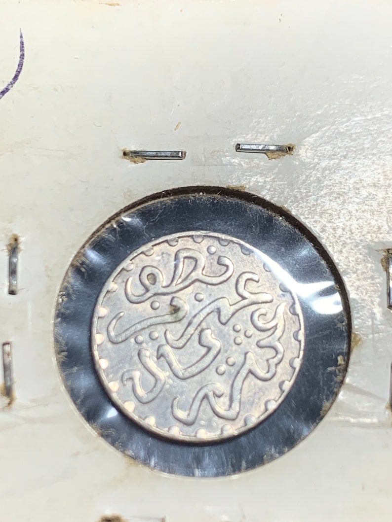 Puede incluir: Una moneda de plata con escritura &aacute;rabe y un dise&ntilde;o decorativo. La moneda est&aacute; encerrada en un soporte de pl&aacute;stico transparente.