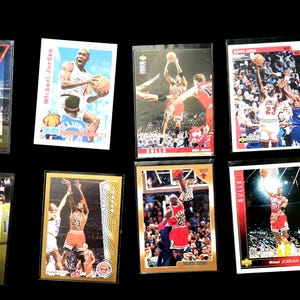 May include: A collection of basketball trading cards featuring action shots of players in various poses. The cards are in protective sleeves and showcase players in red, white, and black uniforms. The background is solid black.