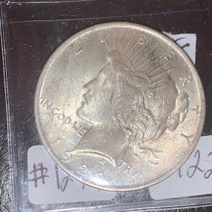 May include: A silver coin with a profile of a woman wearing a radiant crown, inscribed with "IN GOD WE TRUST" and "LIBERTY". The coin is encased in a protective plastic sleeve. The year "1922" is visible.