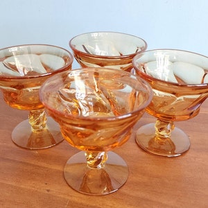 May include: Four amber-colored dessert glasses with a spiral stem and a decorative pattern on the bowl. The glasses are made of glass and are set on a wooden surface.