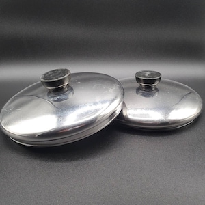 Puede incluir: Dos tapas ovaladas de petaca de acero inoxidable con tapas negras y circulares. Las tapas son reflectantes y muestran un ligero desgaste. El fondo es negro liso.