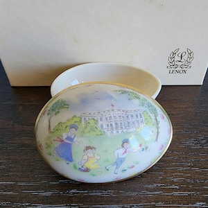 Puede incluir: Caja de porcelana blanca en forma de huevo con ribete dorado. La tapa presenta una escena pintada de niños jugando frente a la Casa Blanca con el texto "LENOX" en la caja.