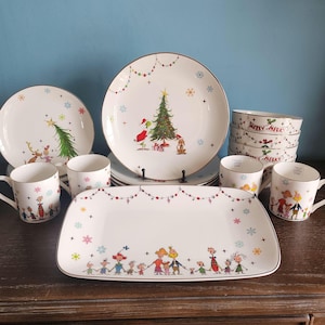 May include: A white ceramic dinnerware set featuring colorful illustrations of characters, Christmas trees, and snowflakes. The collection includes a large rectangular platter, round plates, stacked bowls with Merry Merry text, and mugs with gold-tone rims.