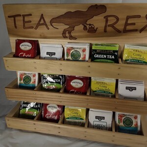 May include: Wooden tea bag organizer with three shelves. The shelves are filled with various tea bags. The top of the organizer has a wooden sign that says "TEA REX" with a dinosaur silhouette.