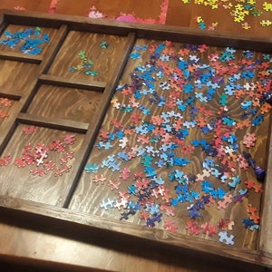 May include: A wooden puzzle tray with a divided section for sorting puzzle pieces. The tray has rope handles and is filled with colorful puzzle pieces.