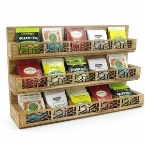 May include: A wooden tea bag organizer with three shelves. The shelves have a decorative cut-out design and are filled with various tea bags. The tea bags are labeled with brand names such as Bigelow and Twinings.