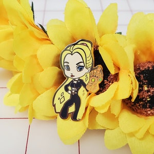 May include: Gold-tone enamel pin featuring a cartoon woman with blonde hair, wearing a black outfit and holding a gold gauntlet with gems. The pin is set against a background of yellow fabric.