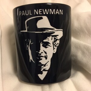 Puede incluir: Taza de cerámica negra con un retrato blanco de Paul Newman con sombrero. El nombre "PAUL NEWMAN" está impreso en blanco encima del retrato. La taza es cilíndrica y está sobre una superficie blanca.