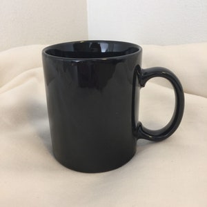 Pode incluir: Uma caneca de caf&eacute; de cer&acirc;mica preta com al&ccedil;a. A caneca est&aacute; sobre uma superf&iacute;cie branca.