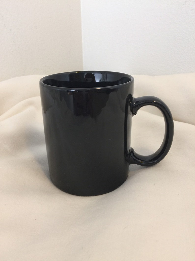 Pode incluir: Uma caneca de caf&eacute; de cer&acirc;mica preta com al&ccedil;a. A caneca est&aacute; sobre uma superf&iacute;cie branca.