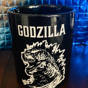 以下が含まれることがあります： 白いゴジラと「GODZILLA」という文字が白い文字で書かれた黒いセラミックマグカップ