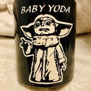 Pode incluir: Caneca de cer&acirc;mica preta com uma imagem branca do Baby Yoda de Star Wars. A caneca tem o texto "BABY YODA" impresso nela.
